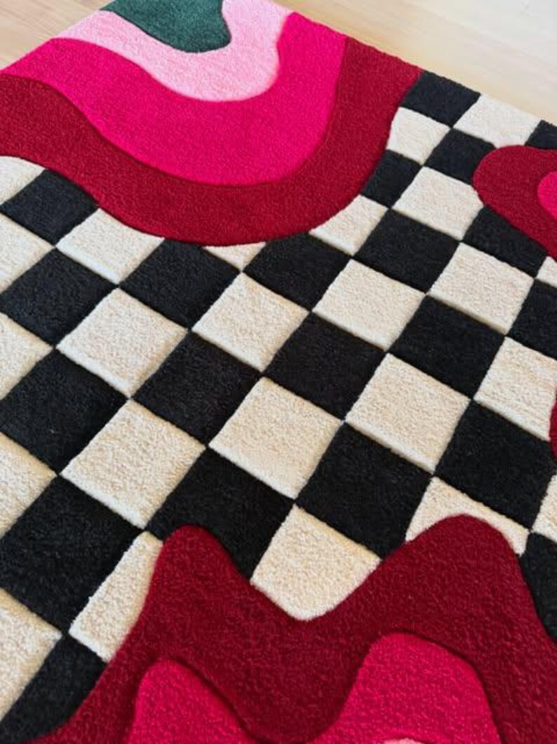 May include: A tufted rug featuring a black and white checkered pattern contrasted with bold, wavy bands of dark red, bright pink, and forest green. The textile surface has a thick, plush pile texture with distinct color-blocked sections.