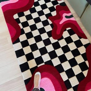 Hand tufted checkerboard rug featuring bold black and white checkered pattern with abstract pink, red, blush and burgundy wavy shapes. Modern handmade wool area rug designed for contemporary living rooms, bedrooms, apartments, dorm rooms and creative interiors. Unique dopamine decor rug with artistic retro aesthetic, colorful statement design and plush texture. Perfect for modern home decor, eclectic interiors, maximalist styling, trendy room makeover, luxury carpet styling, aesthetic room decor