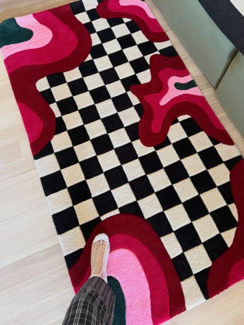 Hand tufted checkerboard rug featuring bold black and white checkered pattern with abstract pink, red, blush and burgundy wavy shapes. Modern handmade wool area rug designed for contemporary living rooms, bedrooms, apartments, dorm rooms and creative interiors. Unique dopamine decor rug with artistic retro aesthetic, colorful statement design and plush texture. Perfect for modern home decor, eclectic interiors, maximalist styling, trendy room makeover, luxury carpet styling, aesthetic room decor