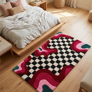 May include: A rectangular area rug featuring a central black and white checkerboard pattern surrounded by abstract, fluid shapes in shades of deep red, bright pink, and dark green. The rug has a tufted texture and a multi-colored, wavy border design.