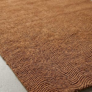 May include: A rectangular area rug in a solid terracotta brown color. The surface of the rug features a consistent, repeating pattern of raised, wavy, horizontal lines that create a textured, rippled effect across the entire fabric surface.