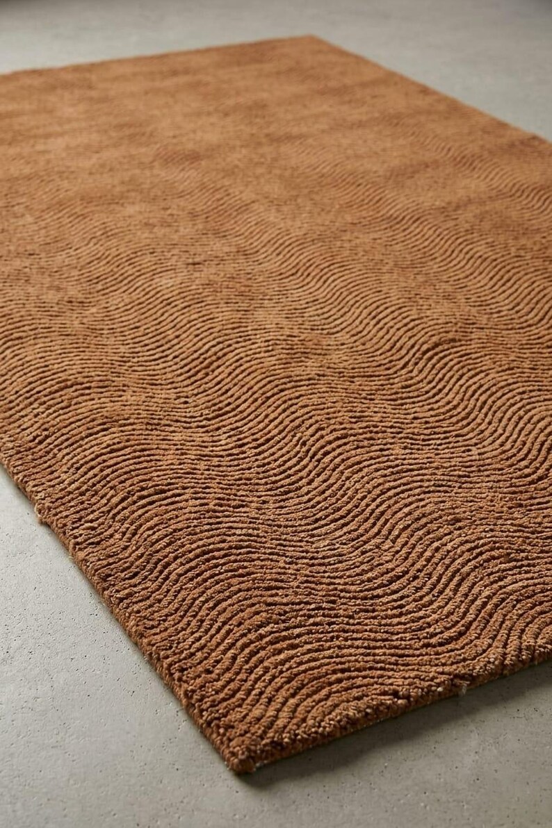 May include: A rectangular area rug in a solid terracotta brown color. The surface of the rug features a consistent, repeating pattern of raised, wavy, horizontal lines that create a textured, rippled effect across the entire fabric surface.
