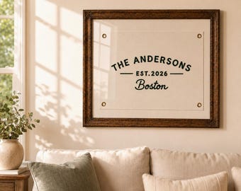 May include: A square wall art piece featuring a cream-colored fabric banner with four brass grommets in a dark brown ornate wooden frame. The text reads THE ANDERSONS, EST. 2026, and Boston in black lettering centered on the fabric banner.
