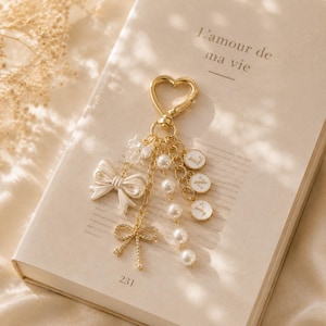 May include: A gold-toned heart-shaped keyring featuring a dangling charm assembly. The charms include a white bow, a smaller rhinestone-encrusted bow, a strand of white pearls, and three circular white discs with the letters L, N, and Y printed in gold.