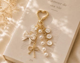 May include: A gold-toned heart-shaped keychain featuring a dangling charm assembly. The charms include a white bow, a smaller rhinestone-encrusted bow, a strand of white pearls, and three circular white discs with the letters L, N, and Y printed in gold.