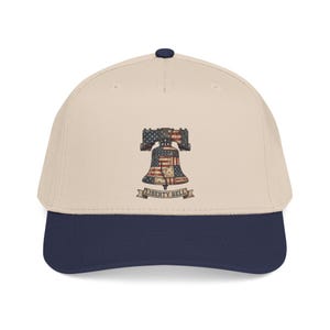 Peut inclure: Une casquette de baseball beige avec une visière bleu foncé et un bouton bleu foncé sur le dessus. L'avant présente un graphique de la Liberty Bell rempli d'un motif de drapeau américain, avec le texte LIBERTY BELL sur une bannière.