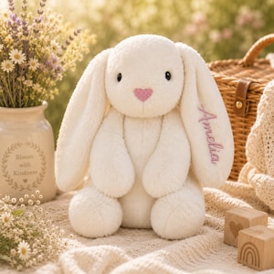 May include: A cream-coloured plush rabbit with long floppy ears, a pink heart-shaped nose, and the name Amelia embroidered in purple script on one ear. The stuffed animal has a soft, fuzzy texture and is seated in an upright position.