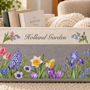 May include: A rectangular storage bin with a cream-coloured top band and a grey base. The cream band features the text "Holland Garden" flanked by floral illustrations. The grey base displays a colourful print of tulips, hyacinths, and irises.
