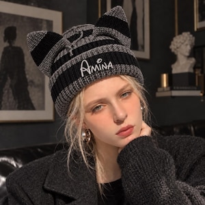 May include: A knit beanie featuring two pointed cat-ear shapes on the top. The hat has horizontal stripes in alternating black and grey colours. The word Amina is embroidered in white script lettering on the front centre of the beanie.