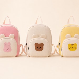 May include: Three small backpacks in pink, cream, and yellow. Each bag features a front pocket with a top handle, two shoulder straps, and a small animal face patch: a pink rabbit, a brown bear, and a yellow tiger, all with rounded ears.