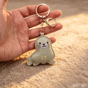May include: A light sage green seal-shaped keyring made of smooth material with stitched edges. The seal features a light grey face with black eyes, a nose, and whiskers. The letters LT are printed in gold on the body, attached to a gold metal ring.