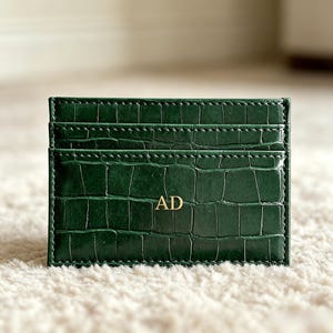 May include: A dark green card holder featuring a textured crocodile skin pattern and visible stitching along the edges. The front of the card holder displays the gold embossed letters AD centered in the middle of the bottom pocket section.