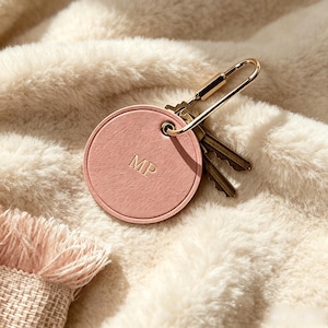 May include: A circular, dusty pink keychain tag with a textured surface and the embossed letters MP in the center. The tag is attached to a gold-toned metal carabiner clip that holds a small cluster of metallic keys, creating a functional accessory.