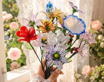 May include: A bouquet of handcrafted glass flowers featuring a vibrant red blossom, a white and blue-rimmed flower, a purple daisy-like bloom, and various other multi-colored glass petals and green stems arranged together in a decorative bunch.