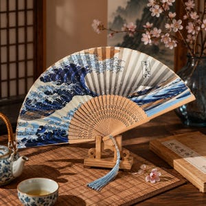 Può includere: Un ventaglio pieghevole con telaio in legno e foglia in tessuto con una stampa grafica blu e bianca di una grande onda oceanica. Il ventaglio è esposto su un piccolo supporto in legno e include una nappa azzurra fissata al perno di base.