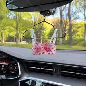 Op de afbeelding: Een miniatuur bralette met een roze en wit hartjespatroon, hangend aan een kleine goudkleurige metalen kledinghanger. Het kledingstuk heeft dunne witte verstelbare bandjes en een geribbelde zoom, hangend aan de binnenspiegel van een voertuig.