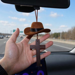 Op de afbeelding: Een bruine vilten cowboyhoed met een zwarte lintband hangend aan een achteruitkijkspiegel, bevestigd aan een donkerbruin houten kruis met een dun bruin koord. De miniatuurhoed en het kruis hangen samen als één auto-accessoire.