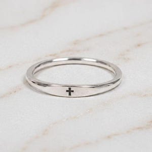 Könnte beinhalten: Ein polierter Ring in Silberoptik mit einer flachen, mittig platzierten rechteckigen Platte, die mit einem kleinen schwarzen Kreuzsymbol graviert ist. Der Ring hat eine glatte, reflektierende Metalloberfläche und ein schmales, rundes Band.