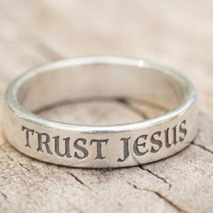 Op de afbeelding: Een zilverkleurige metalen ring met de gegraveerde tekst TRUST JESUS in een schreeflettertype. De ring heeft een gladde, gepolijste afwerking en een consistente breedte, met duidelijke, donker ingekleurde letters op het buitenoppervlak.
