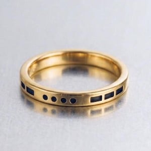 May include: A gold-toned band ring featuring a series of black enamel inlays along the outer surface. The design includes a pattern of small circular dots and rectangular bars arranged horizontally around the circumference of the metal ring.