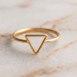 May include: A gold-toned ring featuring a thin, circular band and a prominent, open triangular geometric shape centred on the front. The metal has a smooth, polished finish that reflects light, creating a clean and simple design for this finger accessory.