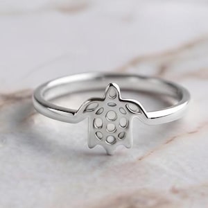 May include: A polished silver-toned ring featuring a central sea turtle motif. The turtle design is integrated into the band and includes cut-out details on the shell, creating a continuous, smooth metal silhouette across the finger.