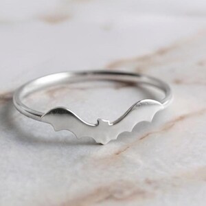 Puede incluir: Un anillo fino de tono plateado pulido que presenta una pequeña silueta de murciélago centrada. La banda es lisa y circular, con el motivo del murciélago integrado directamente en la sección frontal del anillo, creando un diseño metálico.