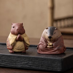 May include: A black ceramic tea cup and two small clay animal figurines on a rectangular black wooden tray. One figurine is a brown capybara with hands pressed together, and the other is a brown beaver-like creature holding a small gold object.