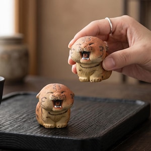 Puede incluir: Dos pequeñas figuras de tigre de cerámica pintadas a mano con acabado mate. Cada tigre tiene un cuerpo redondeado, la boca muy abierta mostrando los dientes y rayas de color marrón oscuro sobre una base de color canela. Una se sostiene.