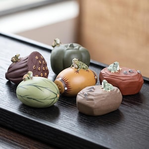 Puede incluir: Una colección de seis pequeñas figuras de cerámica para el té con diversas formas, incluyendo una vaina de loto morada oscura, una calabaza verde y varias piedras lisas, cada una coronada con una pequeña y detallada escultura de rana verde.