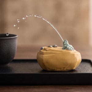 Puede incluir: Un adorno de cerámica amarilla con forma de roca que presenta una pequeña rana verde lanzando un chorro de agua hacia una taza de cerámica gris oscuro. La roca también muestra tres diminutos renacuajos grises sobre su superficie texturizada.