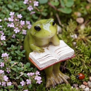Puede incluir: Una pequeña figura de rana verde con grandes ojos negros, apoyando la barbilla en una mano mientras sostiene un libro blanco abierto con páginas visibles. La rana está situada entre pequeñas hojas verdes, diminutas flores moradas y una mariquita roja.