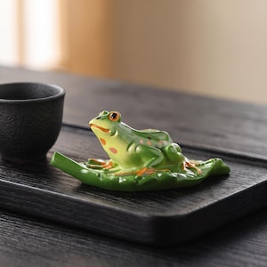 May include: A ceramic frog figurine resting on a green leaf-shaped base, featuring a glossy finish with vibrant green skin, orange-rimmed eyes, and orange spots on its throat and back, positioned next to a small, dark, textured ceramic cup.