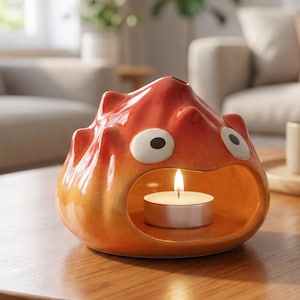 May include: A ceramic candle holder shaped like a stylized flame with a glossy orange and yellow gradient finish. The figure features two large white oval eyes with black pupils and a wide open mouth containing a lit white tea light candle.