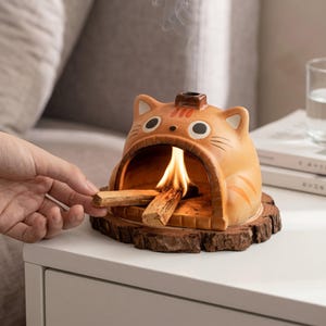 May include: A ceramic incense burner shaped like an orange cat with a wide open mouth, resting on a circular wooden base. The burner contains a burning piece of palo santo wood, with smoke rising from a small chimney on the cat's head.