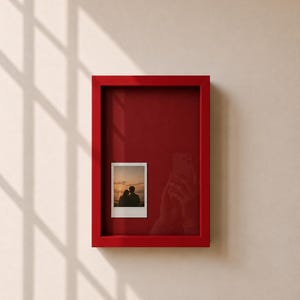 Op de afbeelding: Een rechthoekige rode schaduwdooslijst met een dieprode achterwand. Een enkele verticale instantfoto met een silhouet van twee personen tegen een zonsondergang is bevestigd aan de linkerkant van het binnenoppervlak van de lijst.