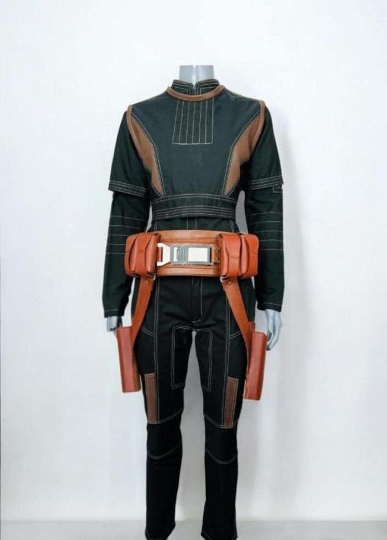 Pu&ograve; includere: Una maglia nera a maniche lunghe e pantaloni abbinati con inserti in similpelle color cuoio e cuciture bianche. Una cintura multiuso color cuoio con due tasche laterali e due attacchi verticali a fondina &egrave; indossata in vita sopra l'indumento.