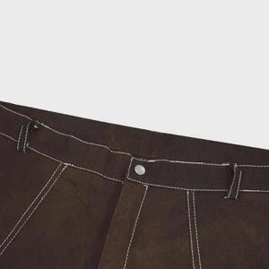 Puede incluir: Pantalones de color marr&oacute;n oscuro con costuras blancas en contraste a lo largo de la cintura, las trabillas para el cintur&oacute;n y las costuras. La prenda incluye un cierre central con bot&oacute;n met&aacute;lico en la cintura y una bragueta estructurada.