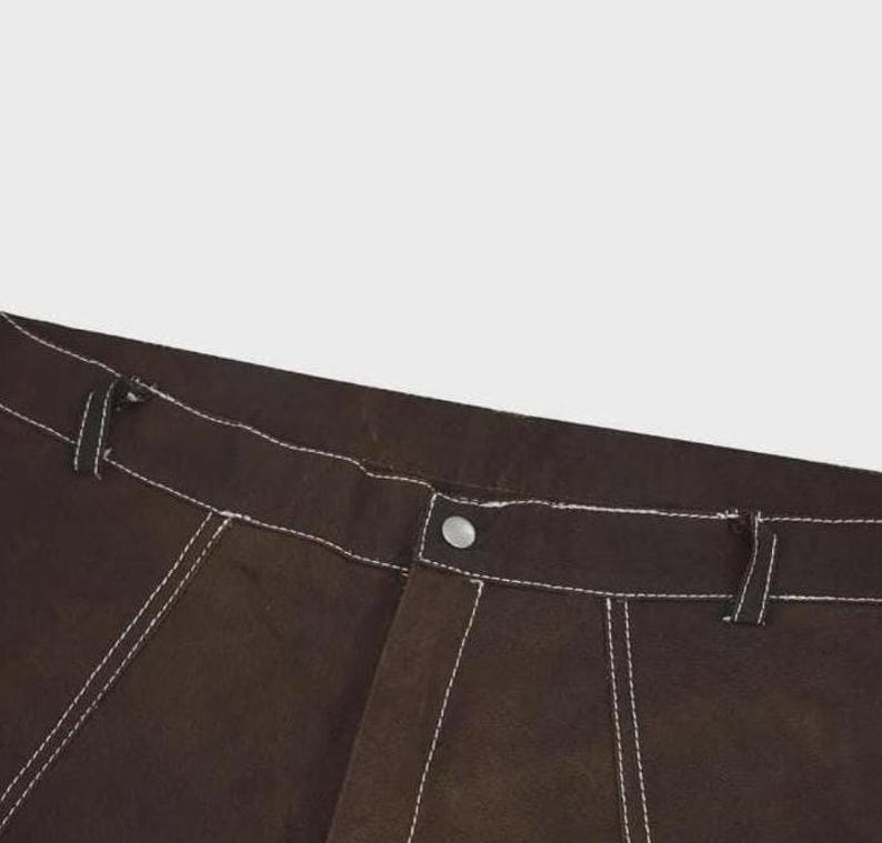 Puede incluir: Pantalones de color marr&oacute;n oscuro con costuras blancas en contraste a lo largo de la cintura, las trabillas para el cintur&oacute;n y las costuras. La prenda incluye un cierre central con bot&oacute;n met&aacute;lico en la cintura y una bragueta estructurada.