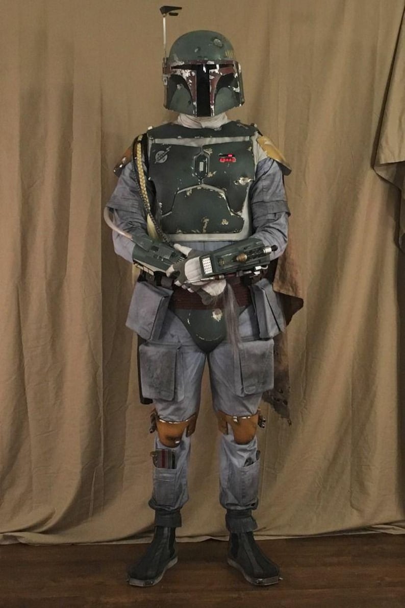 May include: A full-body costume featuring a weathered green helmet with a T-shaped visor and antenna, a dark green chest plate with red accents, grey jumpsuit, utility belt with pouches, gauntlets, and brown boots with a cape attached at the shoulder.