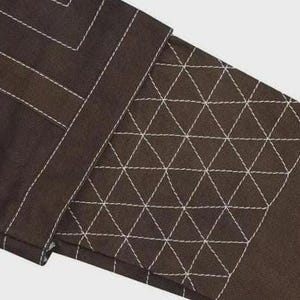 Puede incluir: Una prenda de tela marr&oacute;n oscuro con un prominente patr&oacute;n de cuadr&iacute;cula cosido en blanco. El dise&ntilde;o incluye una secci&oacute;n rectangular con un detalle de costura acolchada triangular densa y repetitiva, contrastada por costuras lineales blancas.
