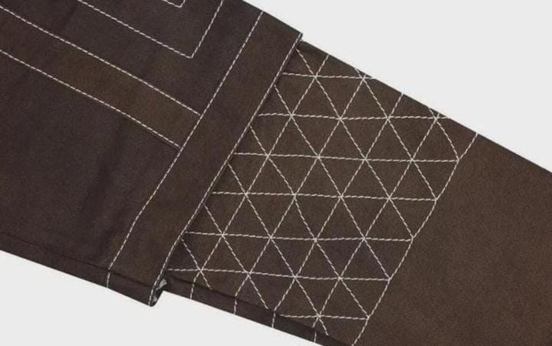 Puede incluir: Una prenda de tela marr&oacute;n oscuro con un prominente patr&oacute;n de cuadr&iacute;cula cosido en blanco. El dise&ntilde;o incluye una secci&oacute;n rectangular con un detalle de costura acolchada triangular densa y repetitiva, contrastada por costuras lineales blancas.