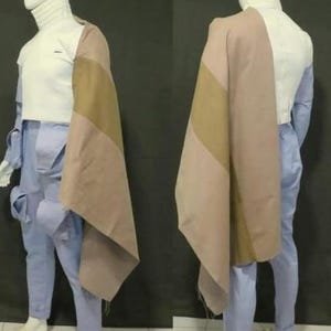 May include: A white turtleneck top paired with light blue cargo pants featuring side pockets and a draped, color-blocked shawl in shades of tan and dusty rose. The ensemble is displayed on a mannequin, showcasing the layered garment construction.