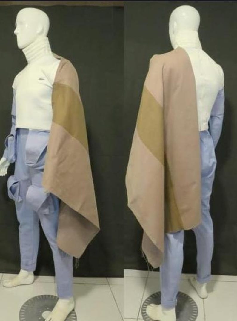 May include: A white turtleneck top paired with light blue cargo pants featuring side pockets and a draped, color-blocked shawl in shades of tan and dusty rose. The ensemble is displayed on a mannequin, showcasing the layered garment construction.