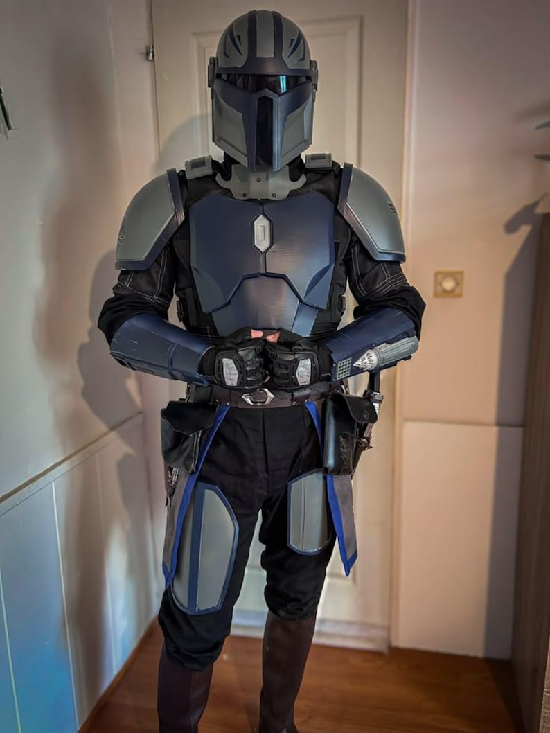Puede incluir: Un traje de armadura completo que presenta un casco con visera en forma de T, hombreras, placa pectoral, guanteletes y protectores de muslos en tonos azul y gris. El traje incluye pantalones negros, guantes oscuros y botas marrones.