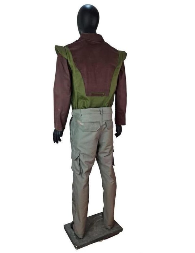 May include: A long-sleeved jacket featuring a dark brown upper section and a green lower panel with extended shoulder flaps, paired with light gray cargo pants. The outfit is displayed on a black mannequin against a plain white background.