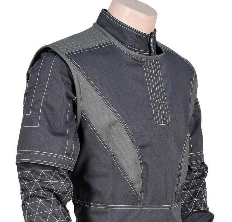 May include: A dark gray tactical-style shirt featuring a high collar, vertical stitched panels on the chest, and reinforced shoulder sections. The sleeves display a white geometric grid pattern, while the torso includes light gray contrast stitching.