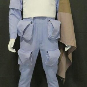 May include: A white high-neck top paired with light blue trousers featuring four large cargo pockets. A tan and brown color-blocked cape drapes over the left shoulder. The outfit is displayed on a white mannequin against a dark background.