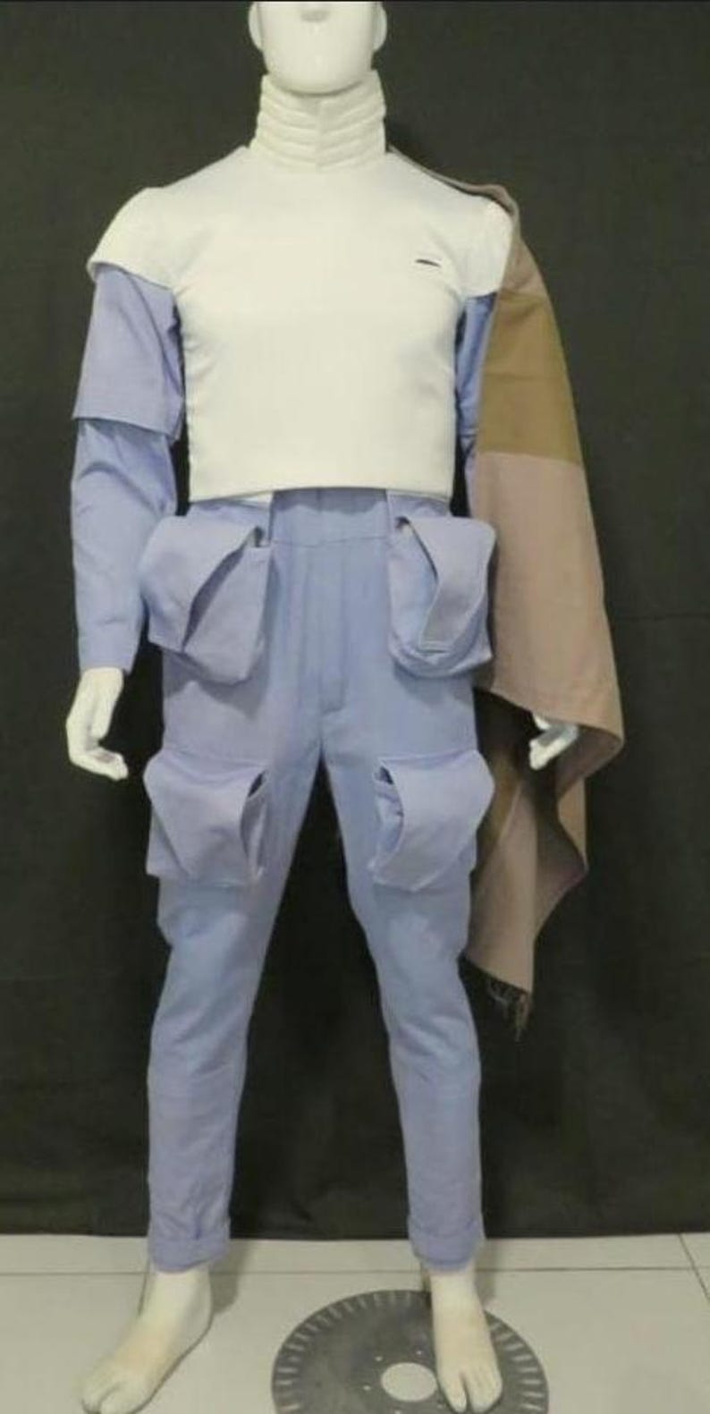 May include: A white high-neck top paired with light blue trousers featuring four large cargo pockets. A tan and brown color-blocked cape drapes over the left shoulder. The outfit is displayed on a white mannequin against a dark background.
