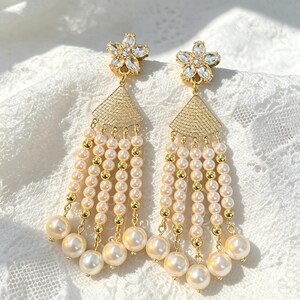 May include: A pair of gold-tone dangle earrings featuring a five-petaled flower stud with clear crystals. Below the stud is a triangular textured gold-tone piece with five cascading strands of cream-colored faux pearls and small gold-tone beads.
